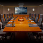 Long boardroom desk centered and surrounded by several chairs in a large board room.